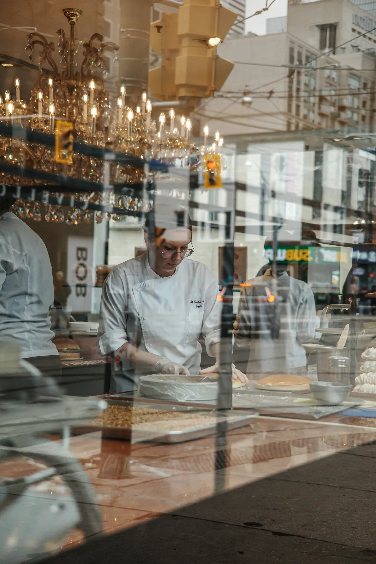 Une pâtissière prépare un gâteau derrière la vitrine de sa boutique, la rue se reflète dans le verre